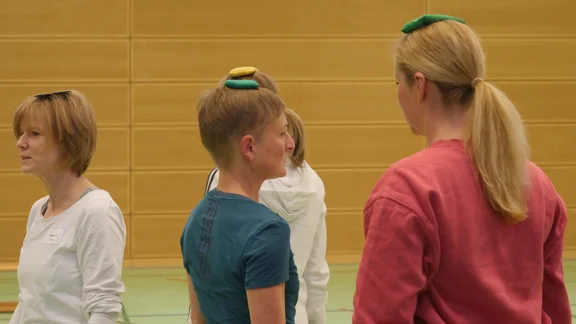 Mehrere Frauen laufen durch die Sporthalle. Auf dem Kopf balancieren sie jeweils einen Bierdeckel oder ein Körnersäckchen.