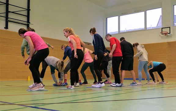 Eine Gruppe Menschen in der Sporthalle bewegen sich auf vor ihnen ausgelegten Teppichfliesen von einer Seite der Halle auf die andere.