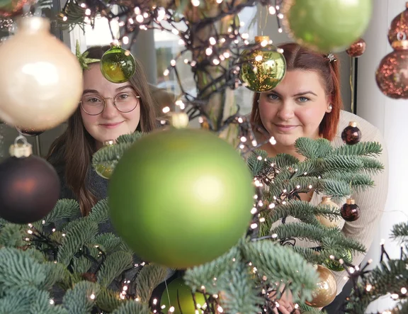 Zwei Auszubildende schauen durch einen geschmückten Weihnachtsbaum lächelnd in die Kamera.