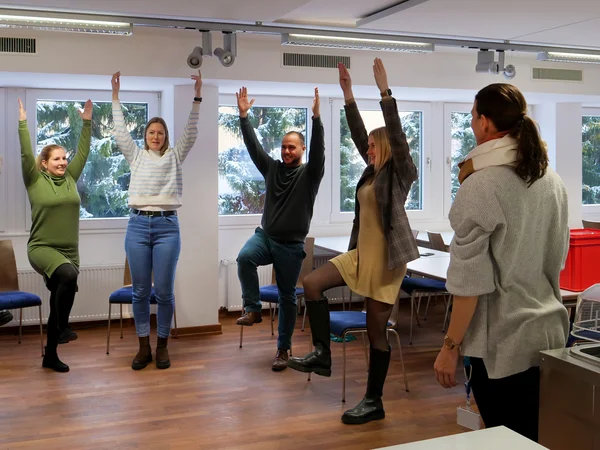 Vier Personen stehen in einem Halbkreis nebeneinander. Sie heben beide Arme zur Decke und stehen auf einem Bein. Eine fünfte Person steht beratend dabei.