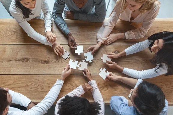 Hände mehrer Personen schieben Puzzleteile auf Tisch zusammen, fröhlich und divers