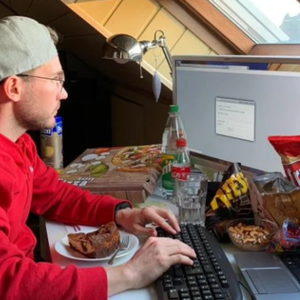 junger Mann an einem chaotischen Schreibtisch arbeitend, der Tisch ist mit ungesundem Essen gefüllt