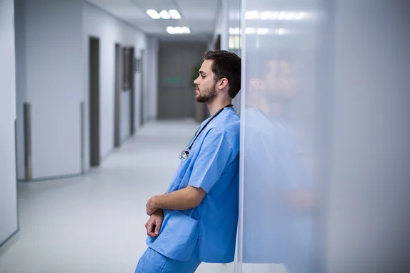 Ein junger Arzt lehnt mit leerem Blick an einer Wand im Krankenhausflur.