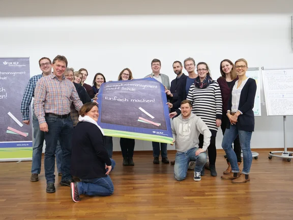 Gruppe von Menschen hält ein Plakat mit der Aufschrift "Gesundheitsmanagement macht Schule. Einfach machen!"