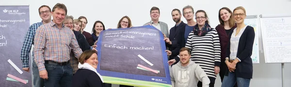 Gruppe von Menschen hält ein Plakat mit der Aufschrift "Gesundheitsmanagement macht Schule. Einfach machen!"