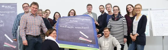 Gruppe von Menschen hält ein Plakat mit der Aufschrift "Gesundheitsmanagement macht Schule. Einfach machen!"