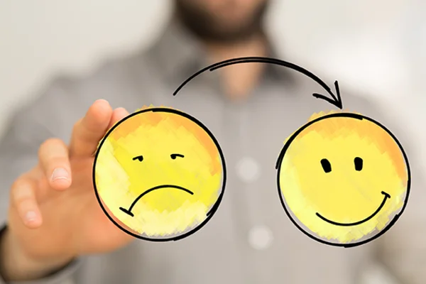 Eine im Hintergrund unscharf abgebildete Person schiebt einen unglücklichen Smiley entlang eines Pfeils zu einem glücklichen Smiley