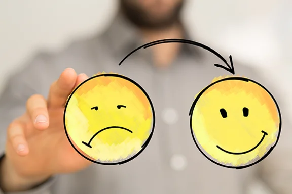 Eine im Hintergrund unscharf abgebildete Person schiebt einen unglücklichen Smiley entlang eines Pfeils zu einem glücklichen Smiley