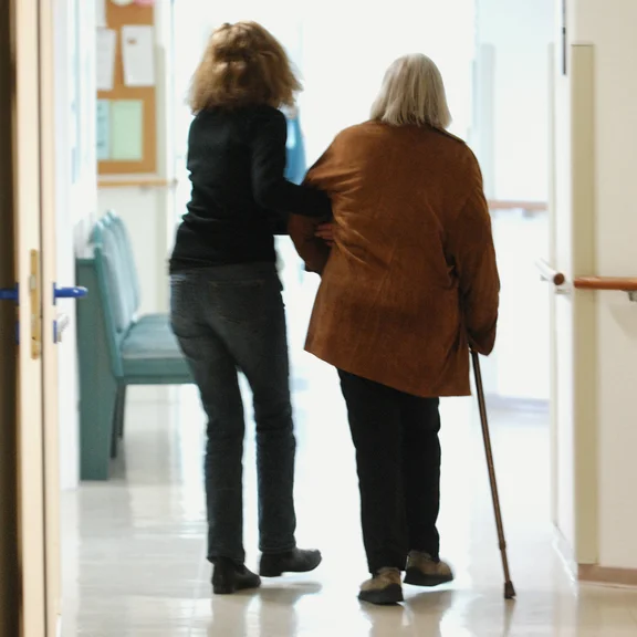 Pflegerin hilft älterer Dame mit Gehstock, indem Sie ihr unter die Arme greift.