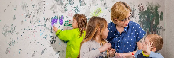 Eine Erzieherin spricht freundlich mit zwei Kita-Kindern. Im Hintergrund malt ein Mädchen mit den Fingern an eine bereits bemalte Wand.