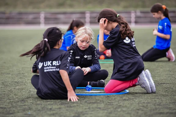 Drei Mädchen sitzen in einem Gymnastikreifen auf dem Sportplatz und schauen auf einen Würfel.