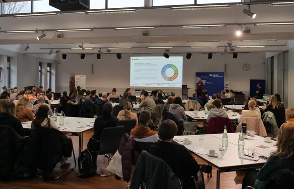 Viele Menschen sitzen (von hinten gesehen) an Tischen und hören einem Vortragenden zu. einem 