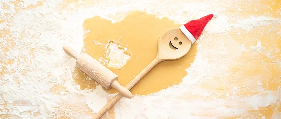 Ein ausgerolltes Teigstück auf einer bemehlten Oberfläche. Aus dem Teigstück ist ein Tannenbaum-Plätzchen ausgestochen. Auf dem Teil liegen ein Nudelholz und ein Holzlöffel mit Gesicht und roter Nikolaus-Mütze.