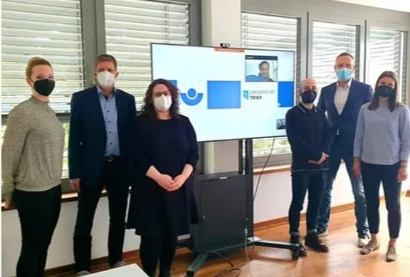 Vertreter von UKRLP und Universität Trier stehen vor Präsentationsbildschirm. Alle tragen Masken.