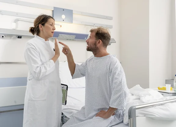Ein Patient sitzt auf dem Krankenhausbett und gestikuliert drohend in Richtung einer Ärztin, die neben dem Bett steht.