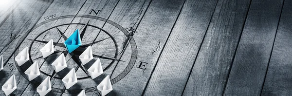 Auf einem grauen Holzboden ist eine Windrose aufgedruckt. Auf dieser Windrose bilden gefaltete weiße Papierschiffchen eine Dreiecksform mit der Spitze nach Norden ausgerichtet. Das Schiffchen an der Spitze ist hellblau.