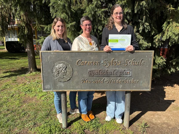 Drei Frauen stehen hinter einem Schild mit der Aufschrift "Carmen-Sylva-Schule".