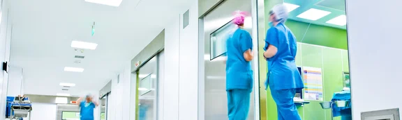Ein Krankenhausflur vor den Operationssälen. Darin steht Krankenhauspersonal mit OP-Häubchen und Mundschutz. 