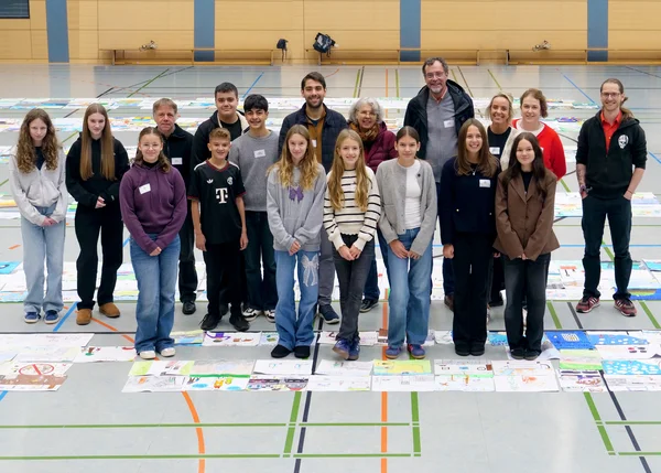 Die Jury des Mal- und Zeichenwettbewerbs steht in der Turnhalle zwischen den eingereichten Bildern.