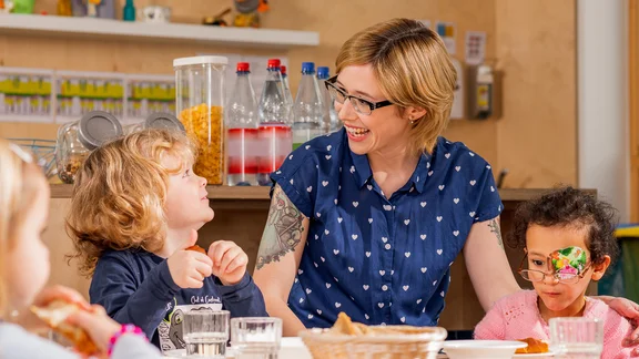 Eine Erzieherin sitzt mit drei Kindern an einem Tisch zum Essen.