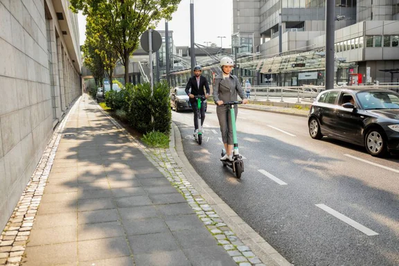 Zwei Personen fahren mit E-Scootern auf einem markierten Fahrradstreifen entlang einer Straße in einer modernen Stadt