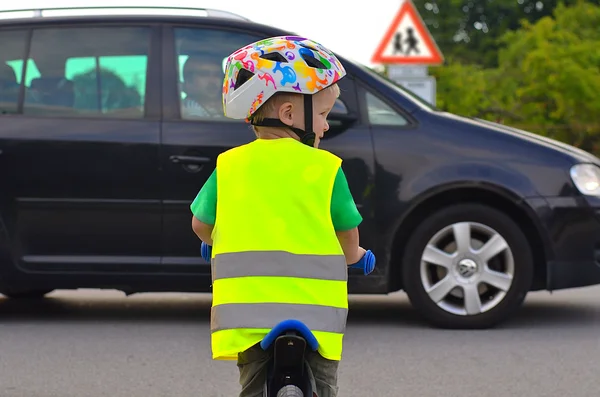 Ein Junge von hinten auf einem Laufrad. Er trägt einen Helm und eine Warnweste und schaut nach rechts. Vor ihm fährt ein Auto von links nach rechts vorbei.