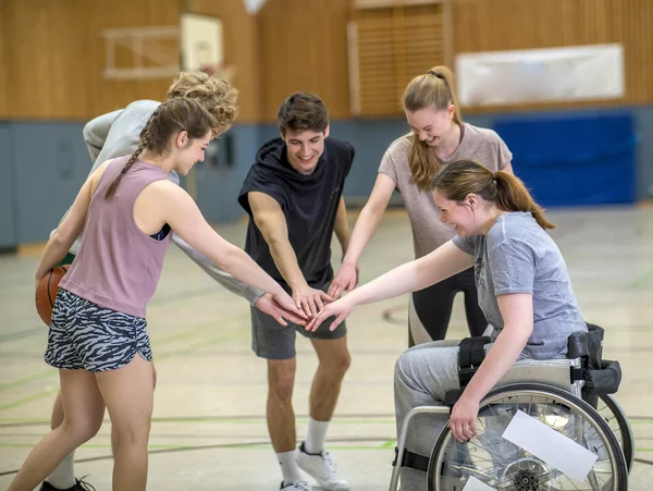 Fünf junge Menschen stehen im Kreis und strecken jeweils eine Hand in die Mitte. Einer dieser Menschen sitzt in einem Rollstuhl.