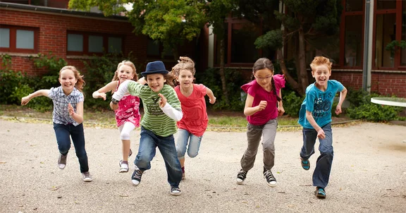 Sechs Schulkinder auf dem Schulhof, im Hintergrund ein Schulgebäude. Sie laufen mit fröhlichen Gesichtern auf den Betrachter zu.