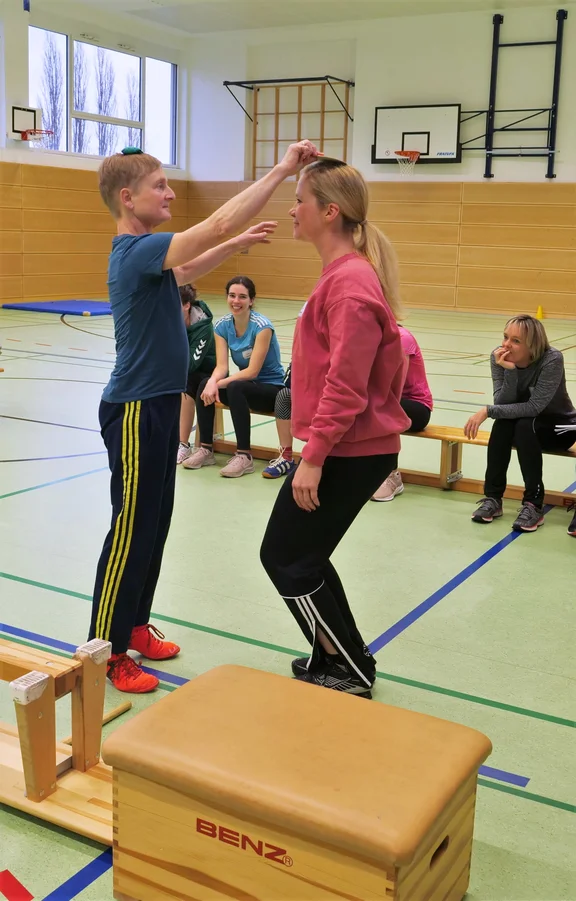 Zwei Frauen stehen sich in der Sporthalle gegenüber. Die eine legt der anderen Frau behutsam einen Bierdeckel auf den Kopf.