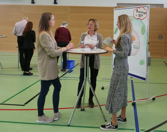Drei Frauen an einem Stehtisch im Gespräch, im Hintergrund eine Flipchart. 