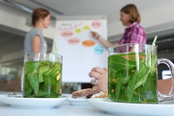 Zwei Tassen Kräutertee im Vordergrund. Verschwommen dahinter stehen zwei Frauen vor einer Flipchart.