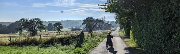 Sabrina Busch fährt Rollstuhl mit Vorspannbike auf einem befestigten Feldweg. Ansicht von hinten auf Sabrina, wie sie der Sonne entgegenfährt. Rechts Wald, links Wiese und Felder.