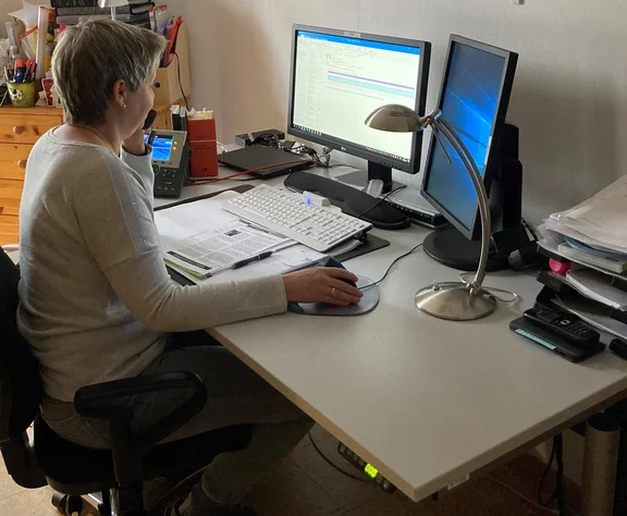 Eine Frau sitzt an ihrem Heimarbeitsplatz und arbeitet. Sie hat eine PC-Maus in der Hand und schaut auf den Bildschirm.
