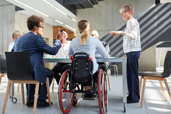 Beschäftigte sitzen und stehen rund um einen Besprechungstisch, darunter eine Frau, die in einem Rollstuhl sitzt.