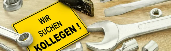 Mehrere Werkzeuge liegen auf einem Tisch. Mittendrin ein gelbes Schild mit der Aufschrift "Wir suchen Kollegen!"