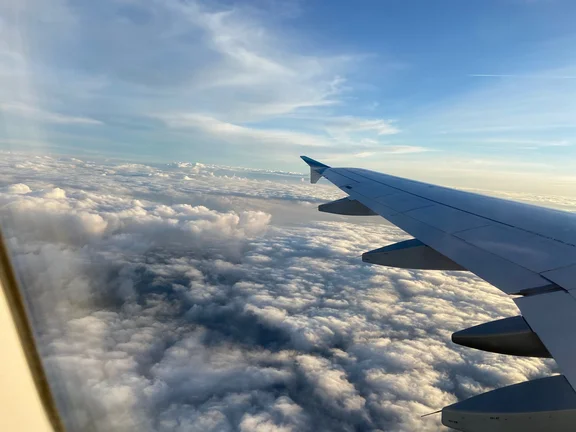 Blick aus dem Fenster eines Flugzeugs auf den Flügel, darunter weiße Wolken im Sonnenschein.