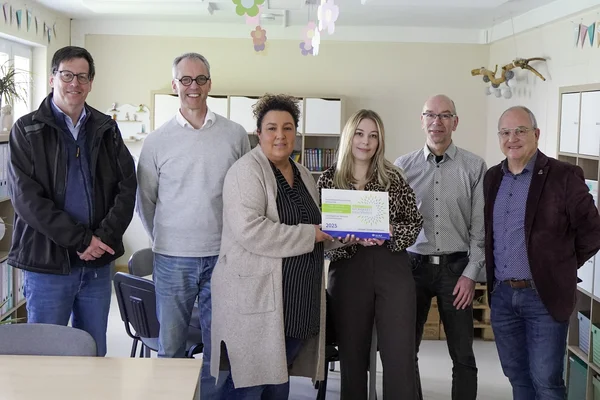 Gruppenfoto in einem Klassenraum: Sechs Erwachsene stehen zusammen, in der Mitte wird die Präventionspreisplakette „Sicherheit und Gesundheit“ für 2025 in die Kamera gehalten.