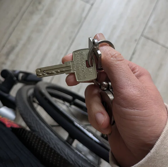 Eine Frau fotografiert einen EU-Toilettenschlüssel in ihrer Hand. Dabei ist auch ein Anschnitt ihres Rollstuhl zu sehen.