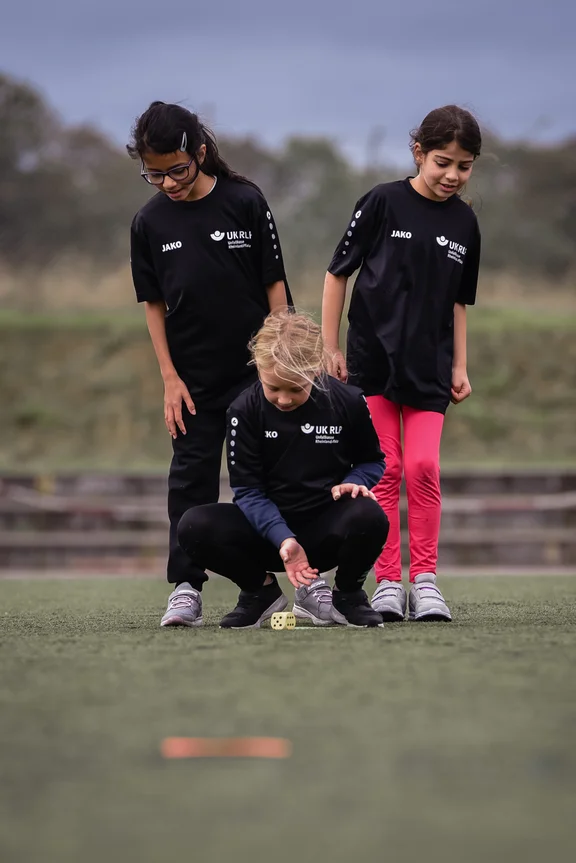 Drei Mädchen auf dem Sportplatz. Eins davon hockt und wirft einen Würfel. Die anderen schauen ihm über die Schulter.