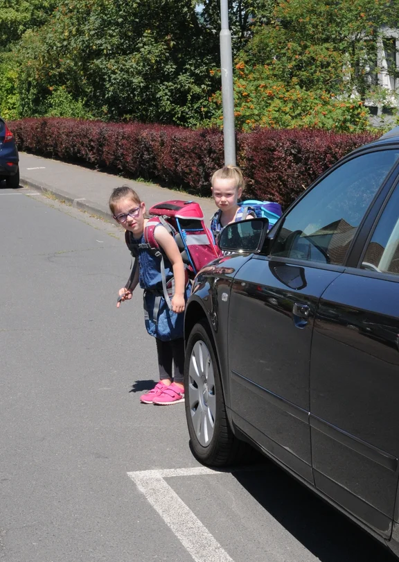Zwei Kinder schauen hinter einem parkenden Auto auf die Straße. 