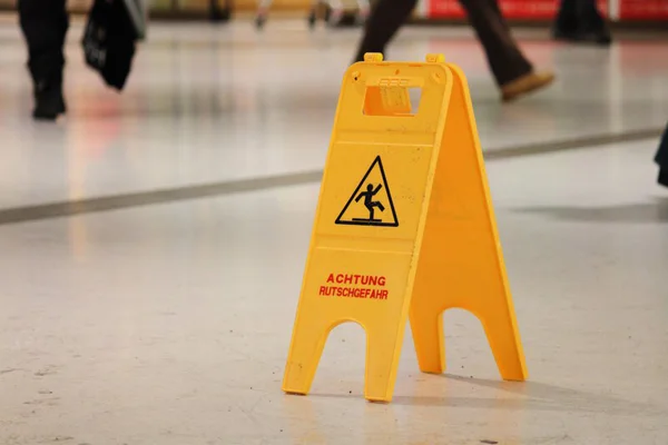 Ein gelbes Warnschild mit der Aufschrift 'ACHTUNG RUTSCHGEFAHR' steht auf einem glänzenden Boden in einem belebten Innenbereich.