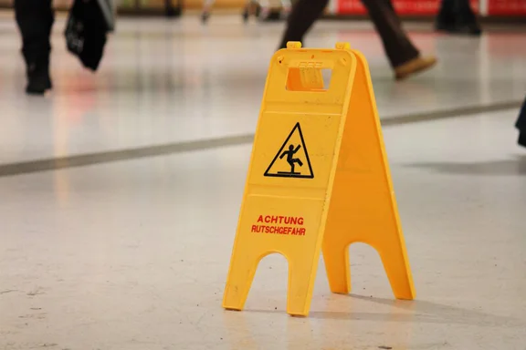 Ein gelbes Warnschild mit der Aufschrift 'ACHTUNG RUTSCHGEFAHR' steht auf einem glänzenden Boden in einem belebten Innenbereich.