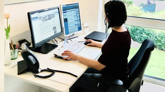 Frau sitzt im Homeoffice am Schreibtisch, vor 2 Monitoren. Auf dem Schreibtisch steht ein kabelgebundenes Telefon und sie trägt ein Headset.