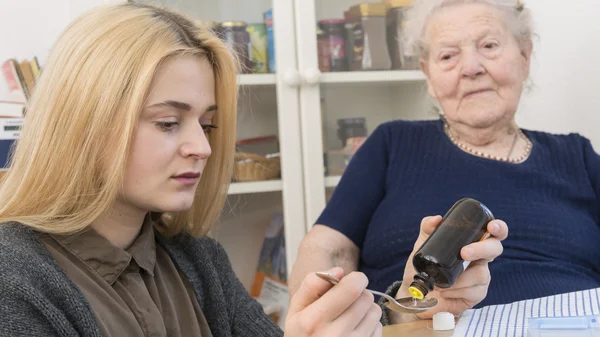 Junge Frau gibt medizinische Tropfen auf einen Löffe, sitzend am Tisch. Im Hintergrund sieht ihr eine ältere Dame zu.l