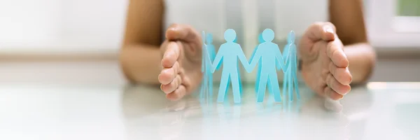 Aus Papier ausgeschnitteten Menschen halten sich im Kreis stehend an den Händen. Zwei menschliche Hände schützen sie von den Seiten.