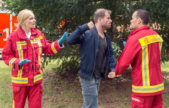 Ein aggressiver Mann droht mit erhobener Faust einem Rettungssanitäter. Eine zweite Rettungssanitäterin versucht den Mann zu beruhigen.