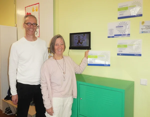 Ein Mann und eine Frau stehen vor einer Wand, an der mehrere Präventionspreis-Plaketten aufgehängt sind.