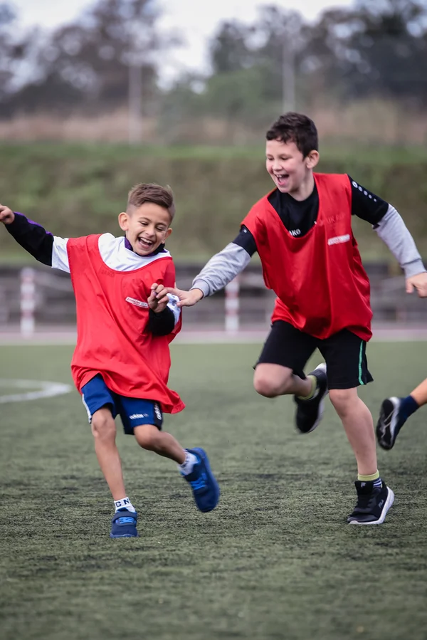 Zwei lachende Kinder in Sportkleidung toben auf dem Sportplatz.